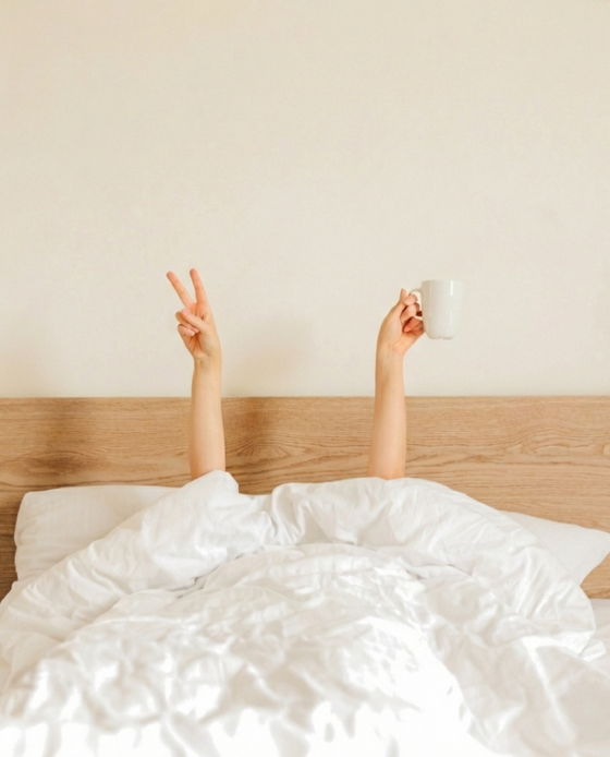 Frau liegt mit Bettdecke über Kopf im Bett und streckt ihre Hände nach oben. In einer Hand hält sie eine Tasse